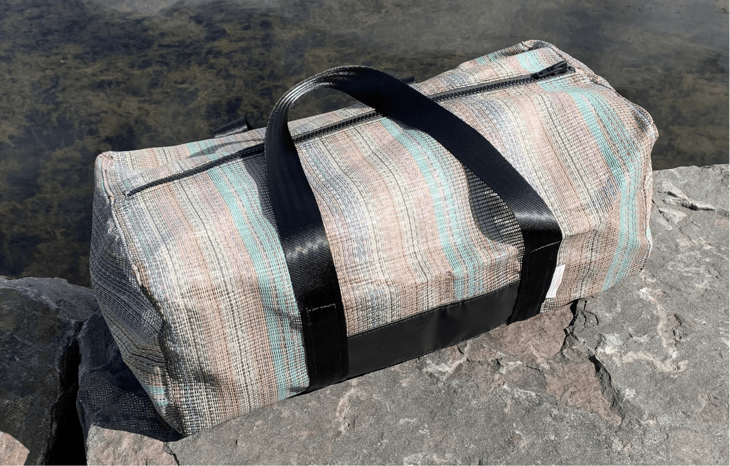 Rainbow-patterned, upcycled sports bag duffel sitting on rocks beside a pond.