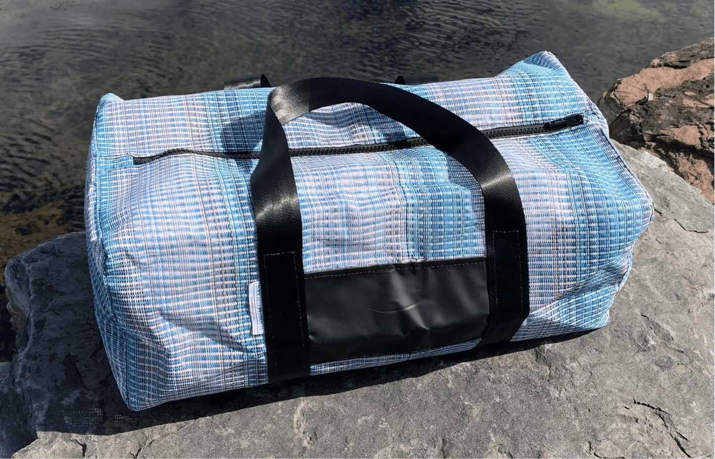 Rainbow-patterned, medium-sized sports bag sitting on rocks beside a pond.