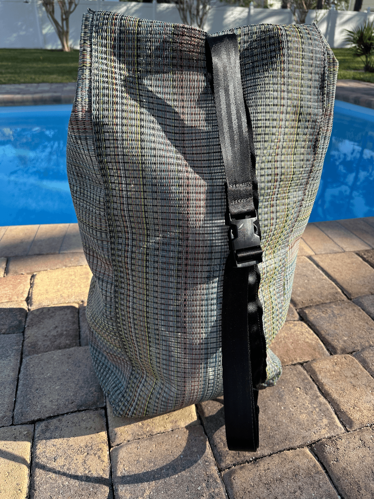 Rainbow mesh sports bag sitting on brick in front of a swimming pool.